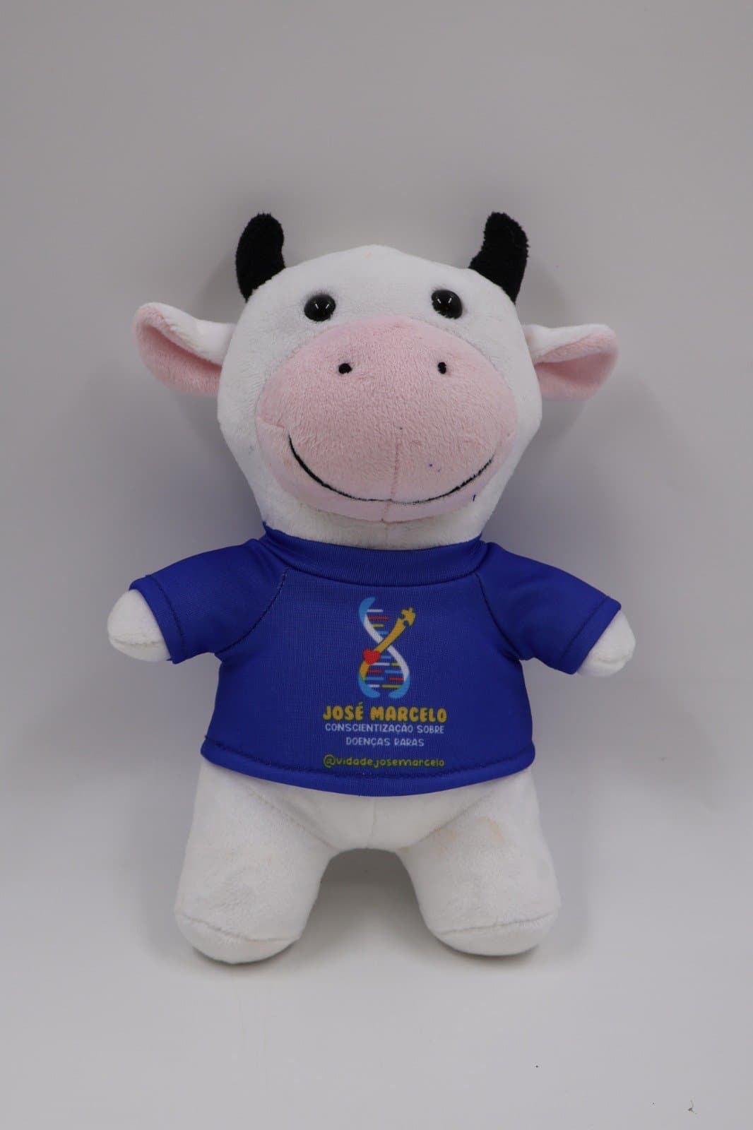 Vaquinha do Zé, pelúcia branca com chifres pretos e focinho rosa, vestindo camiseta azul com o símbolo da campanha — vista de frente